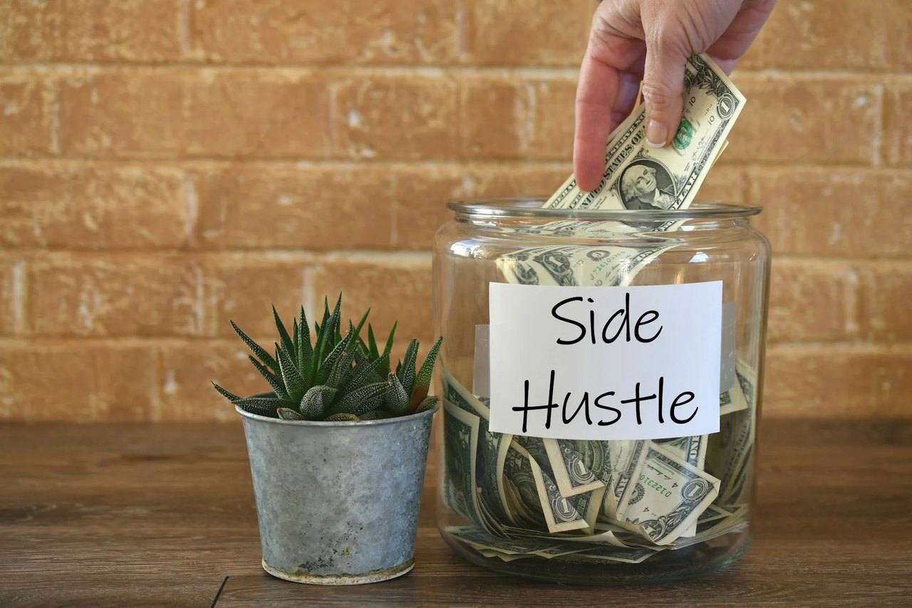 Hand legt Dollarnote in Glas mit "Side Hustle"-Etikett, gefüllt mit Geld, daneben eine Pflanze.