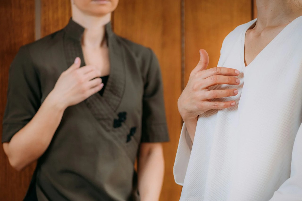 Zwei Frauen mit Händen auf der Brust, eine in olivgrüner, die andere in weißer Kleidung vor Holzpaneel.