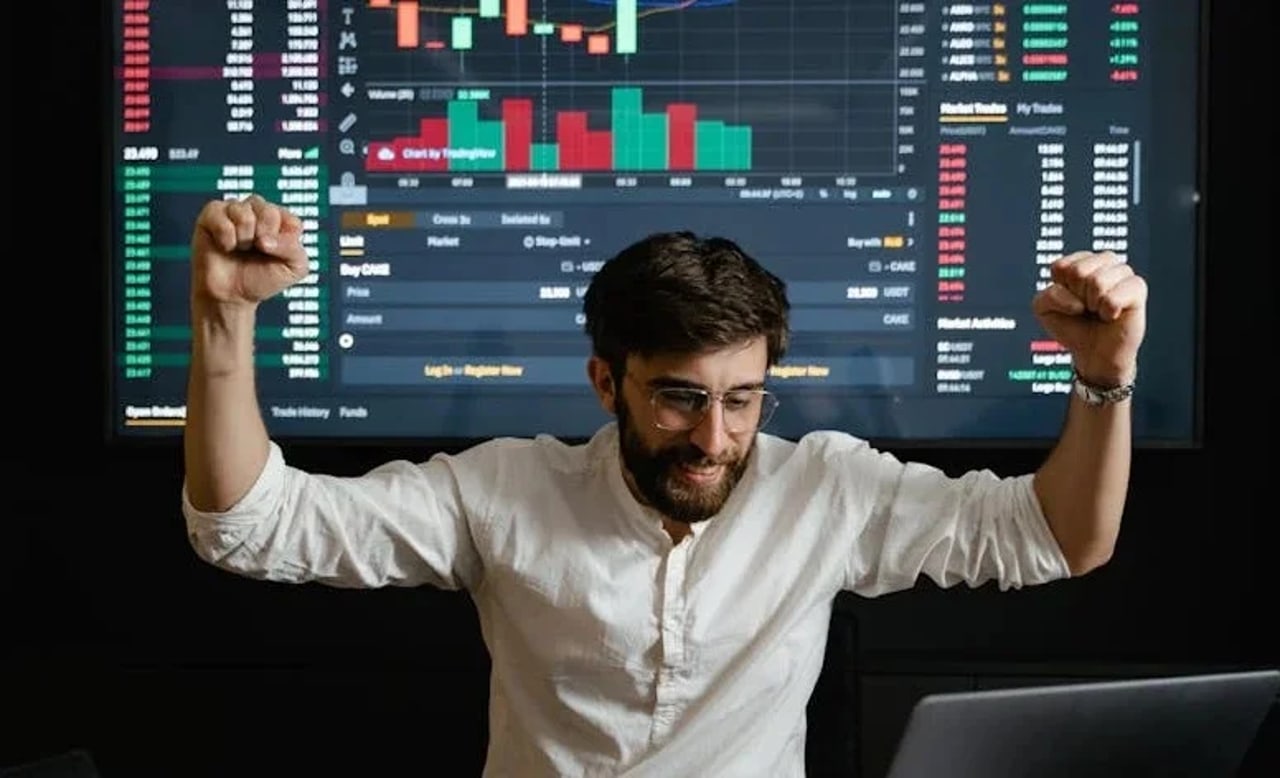 Ein Mann feiert mit erhobenen Fäusten vor einem Bildschirm mit Finanzdiagrammen und -daten.