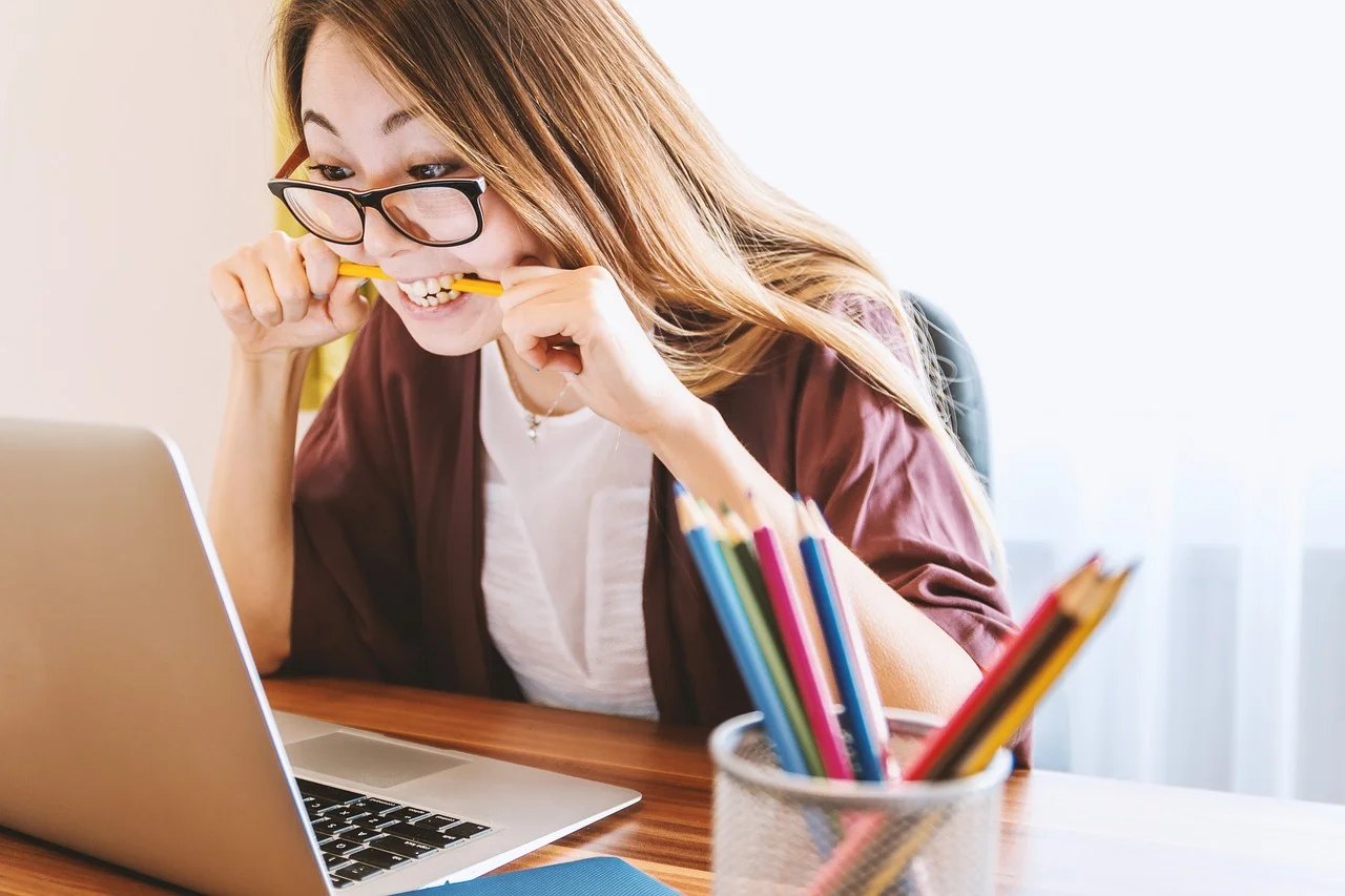 Junge Frau mit Brille kaut nervös auf einem Bleistift, während sie auf einen Laptop schaut.